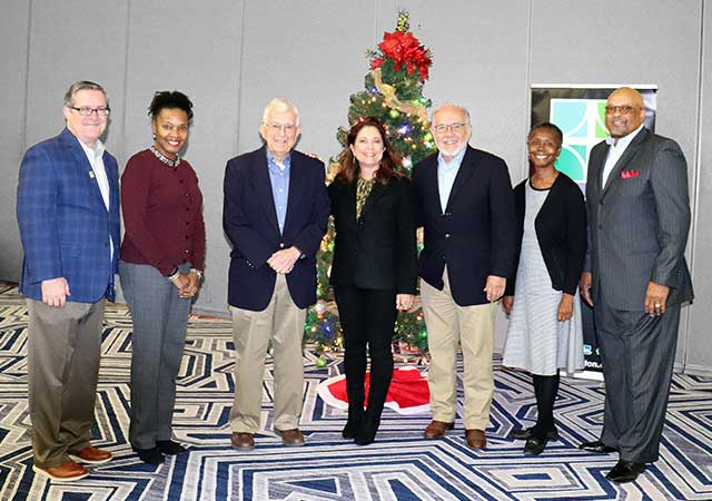 Karen Marshall stands with a group of six colleagues in front of a decorated Christmas tree. The group is dressed in business casual attire and smiles warmly for the photo.