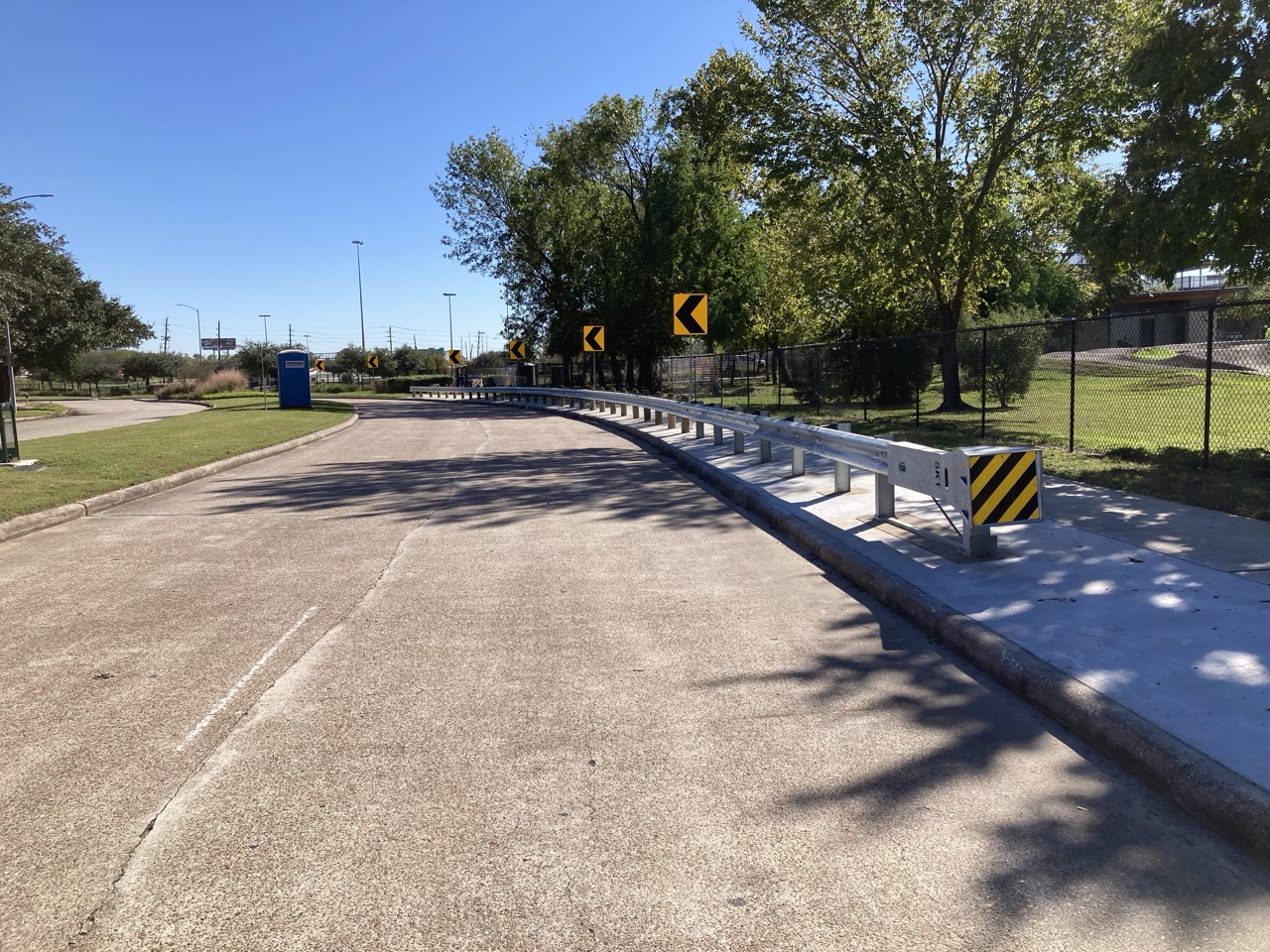 A newly installed metal guardrail lines a curved roadway under a clear blue sky. Yellow directional arrow signs guide traffic, and trees and a chain-link fence border the road. A portable toilet is visible on a grassy area.