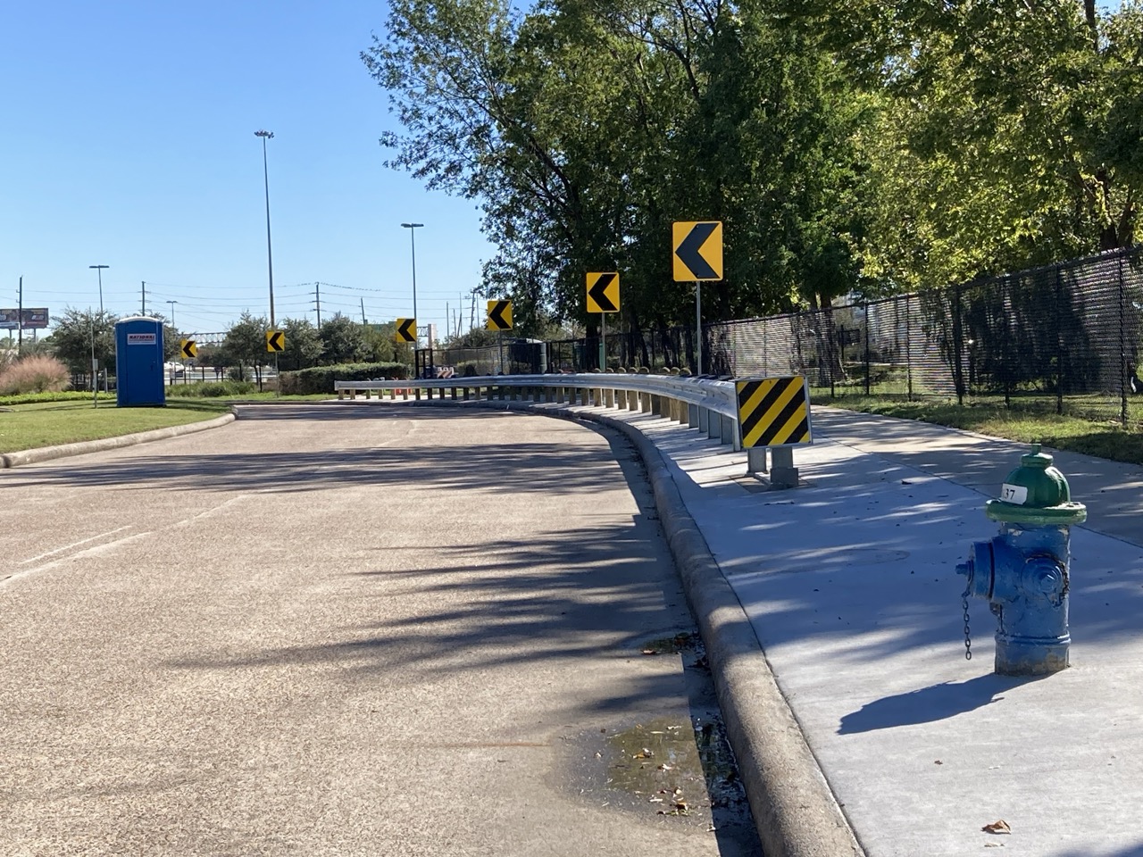 A metal guardrail follows the curve of the road with directional arrow signs. A blue fire hydrant sits near the curb, and a chain-link fence borders the area. Trees and clear skies complete the scene.