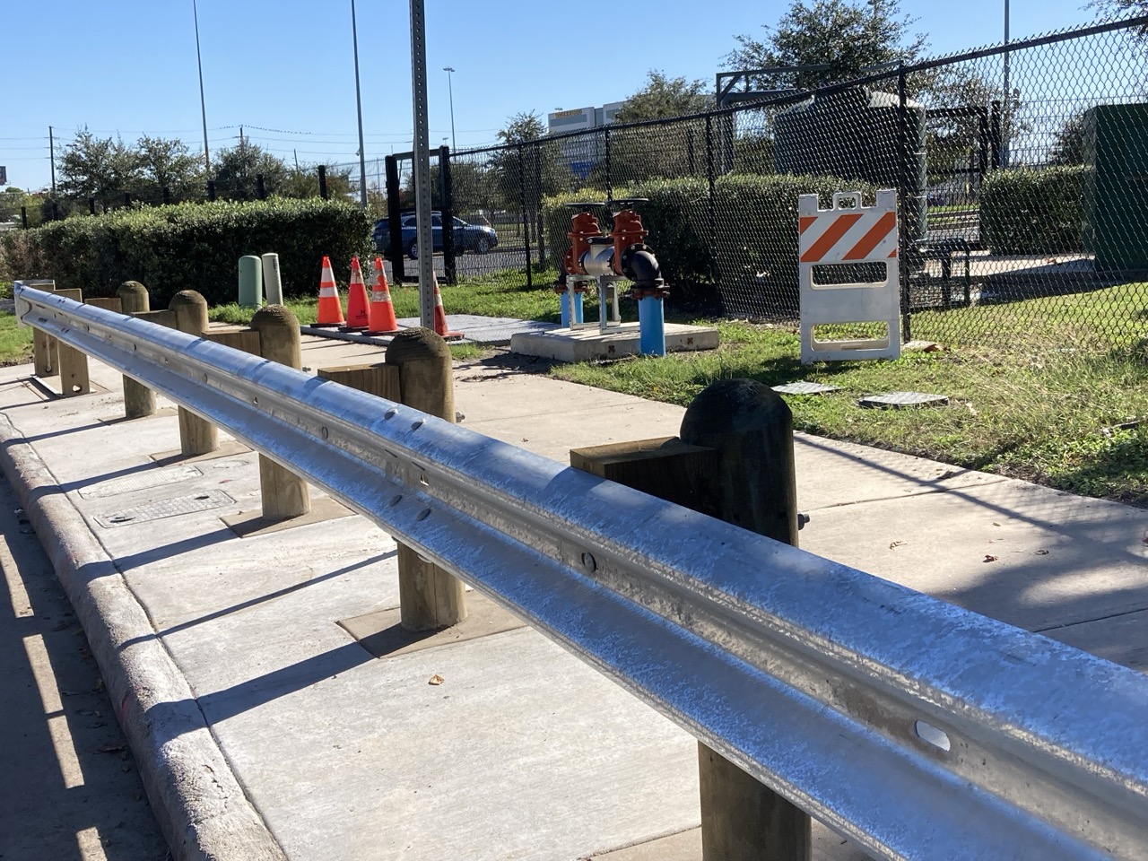 A close-up of a metal guardrail along a sidewalk shows wood posts securing it. Behind the fence, orange cones, utility pipes, and a striped barricade are visible. A chain-link fence and shrubs line the background.