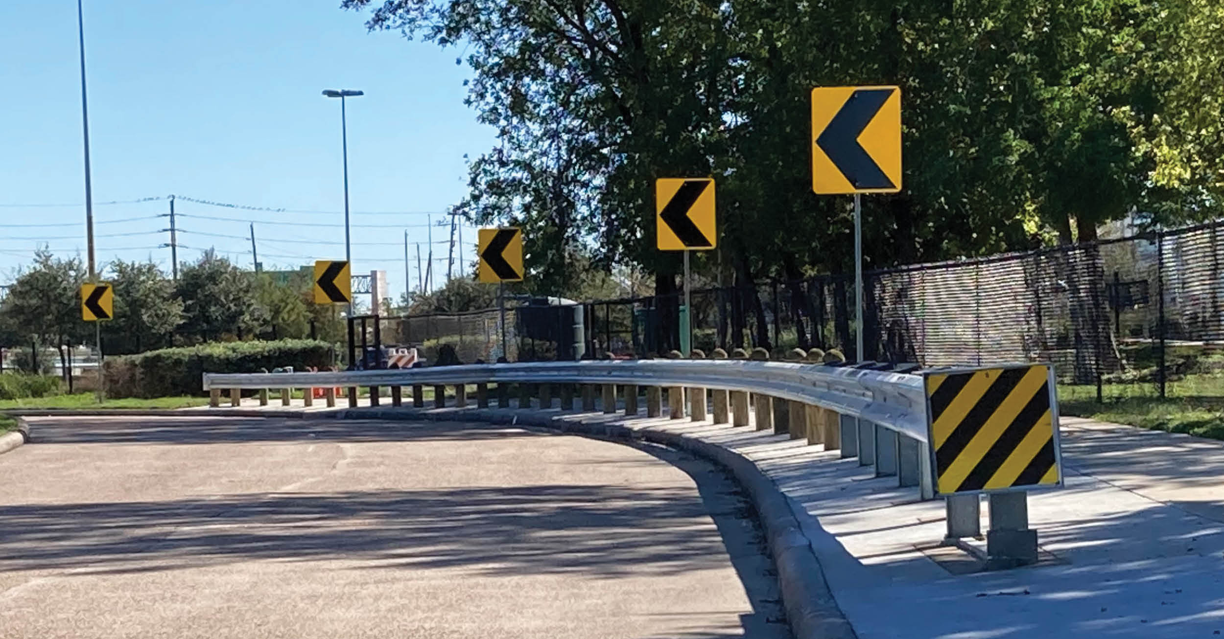 A metal guardrail follows the curve of the road with directional arrow signs. A blue fire hydrant sits near the curb, and a chain-link fence borders the area. Trees and clear skies complete the scene.