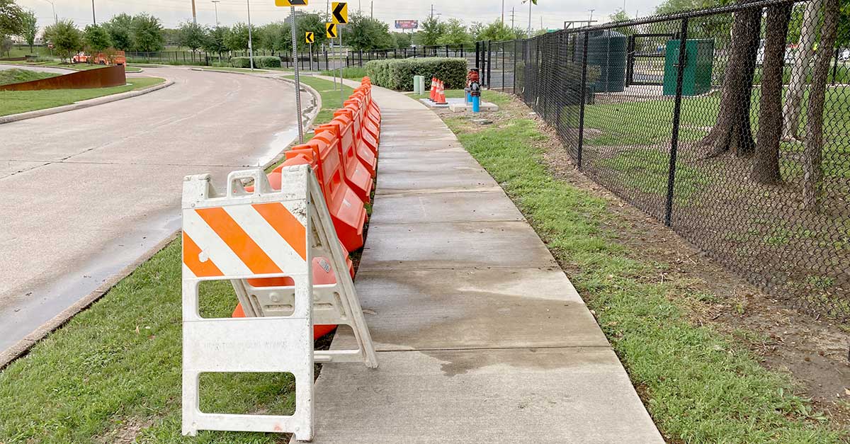 A sidewalk curves alongside a road, lined with orange traffic barriers and a chain-link fence on the right. A white barricade sits in the foreground.