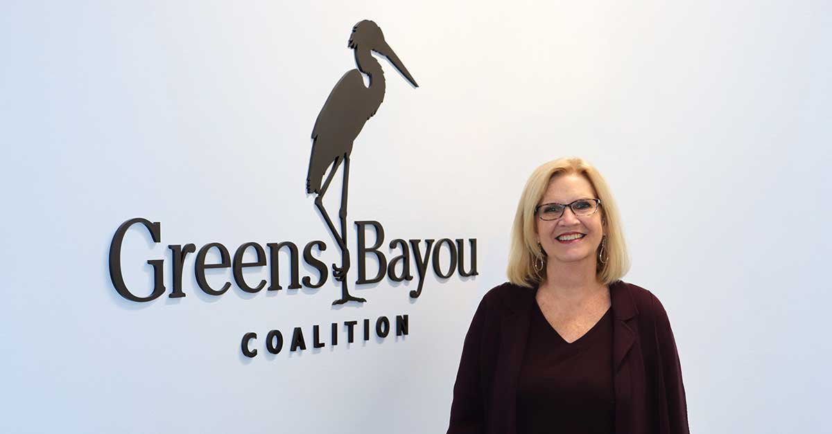 A woman stands smiling in front of a wall featuring the logo of the "Greens Bayou Coalition." The logo includes a silhouette of a heron, standing on one leg. The woman is wearing glasses, a dark top, and a dark jacket. The background is light, drawing attention to the logo and the woman.