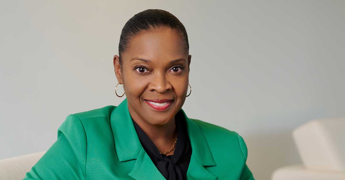 This image features a professional portrait of a woman with a warm, confident smile. She is wearing a green blazer over a black blouse, which is tied at the neck in a subtle bow. Her hair is neatly pulled back, accentuating her face. She wears small gold hoop earrings and has a natural, polished makeup look. The background is softly blurred in neutral tones, keeping the focus on her expression and attire. The overall setting appears clean and professional, enhancing her poised and approachable demeanor.