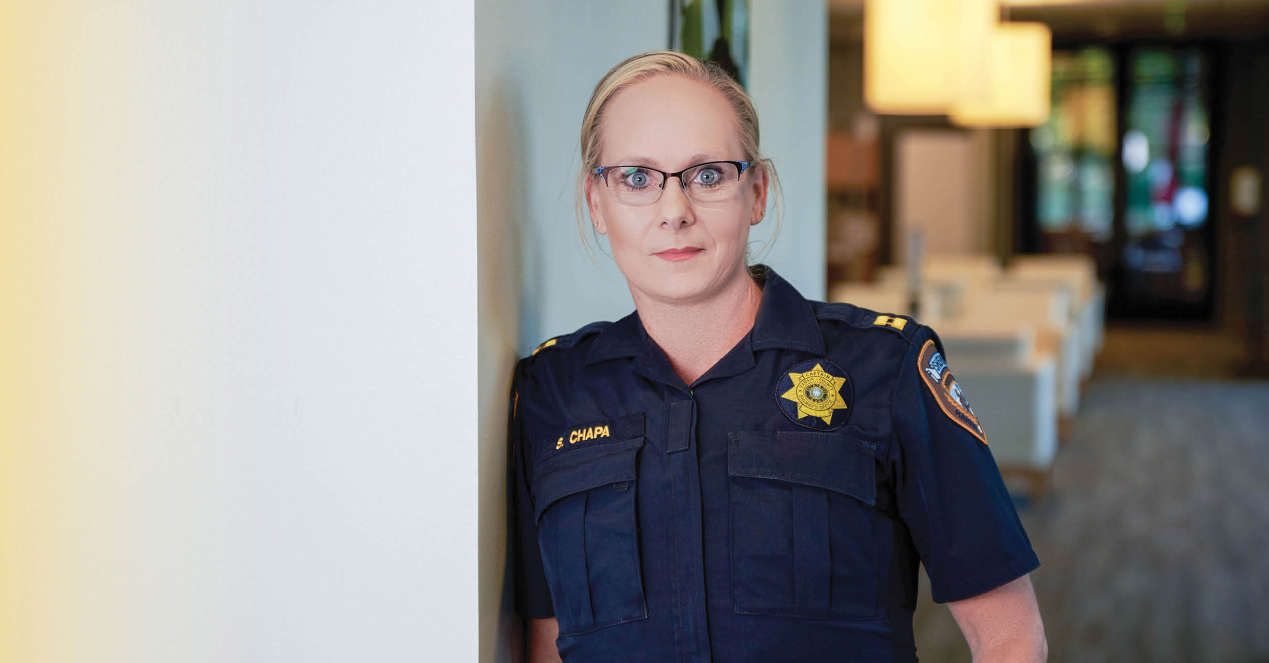 Close-up of Sandi Chapa in uniform, leaning against a wall indoors. The background shows a blurred modern office or meeting area with soft lighting.