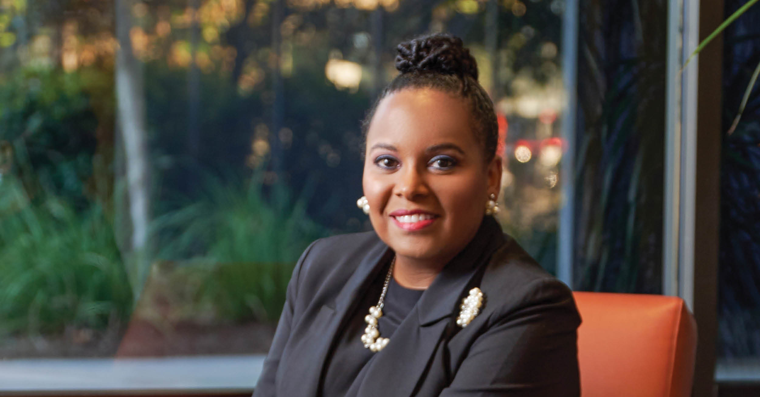 A professional headshot of Sorita Alexander sitting in an orange chair, wearing a black blazer with pearl accessories. She smiles warmly with greenery visible through a window behind her.