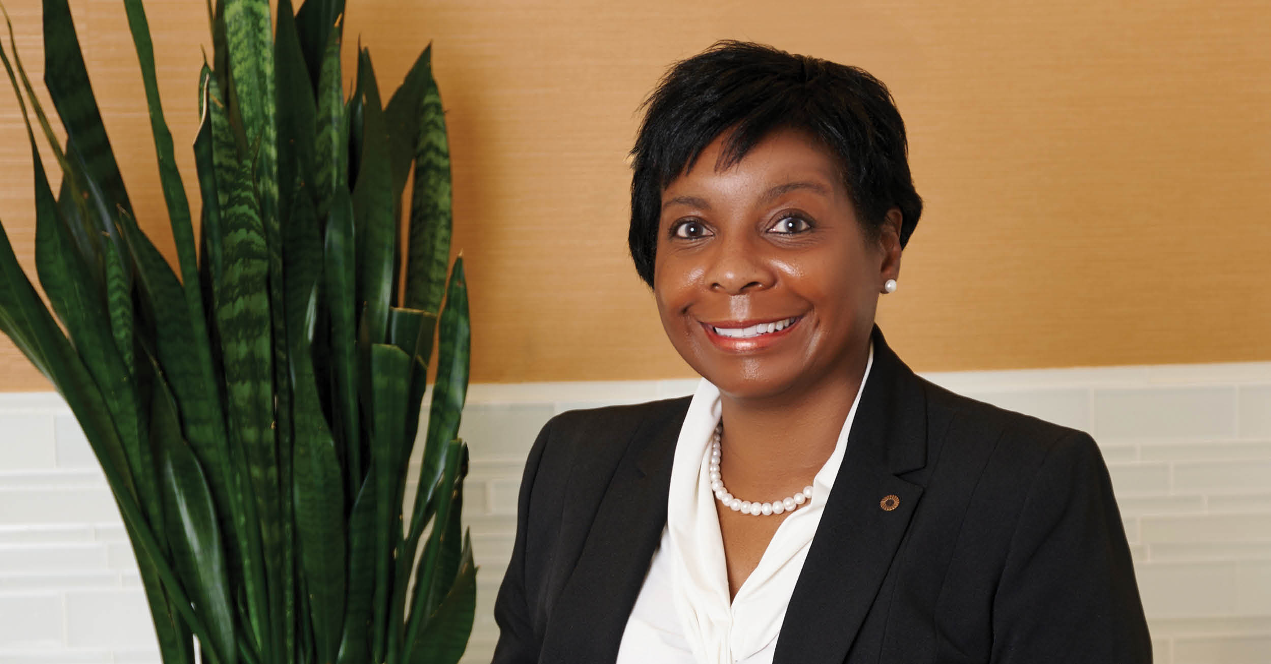 Tina Poindexter smiles in a business portrait, wearing a dark blazer and pearl necklace, standing beside a tall green plant against a warm-toned wall featured as a North Houston Leading Lady.
