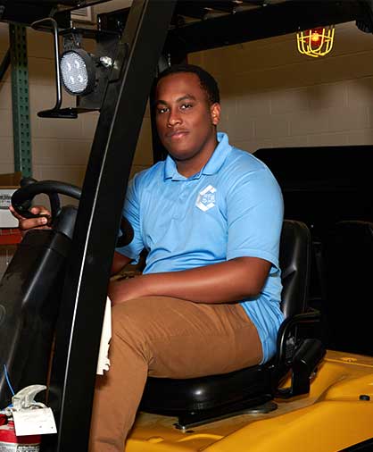 Logistics & Distribution Program 2 Close-up of a student in a light blue polo sitting confidently in a forklift, surrounded by warehouse shelving.