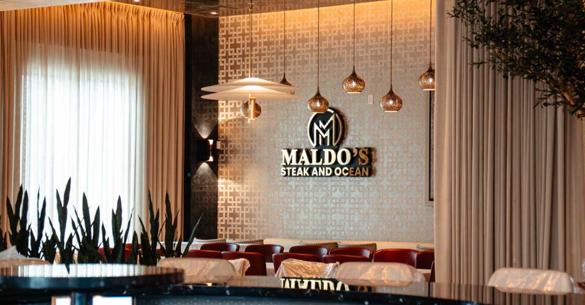 Maldos Newsroom Graphic Interior view of Maldo’s Steak and Ocean restaurant showing elegant lighting, tall curtains, and the restaurant logo on a patterned wall behind red seating.