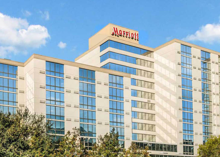 A bright daytime photo of the Houston Marriott North hotel featuring its modern glass windows, cream-colored exterior, and blue sky with scattered clouds overhead.