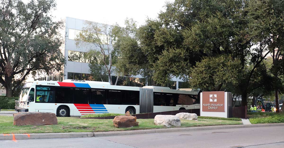 A METRO bus passes by a North Houston District monument sign, surrounded by landscaped greenery and mature trees, with a sleek modern office building in the background.