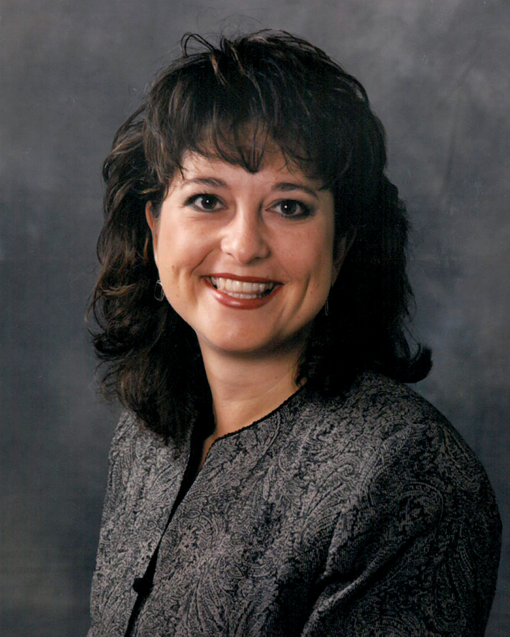 A portrait of a smiling woman with shoulder-length dark brown hair styled in soft curls. She wears a dark, patterned blazer against a solid dark gray background.