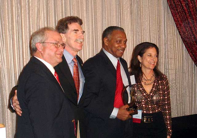 Four individuals in formal attire smile for a group photo. One man holds an award while standing with two other men and a woman. A backdrop with light curtains and red drapes frames the setting.