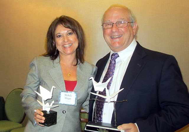 A smiling woman and a man hold matching white bird-shaped awards on black bases. Both are dressed in business attire and stand close together against a beige wall background.