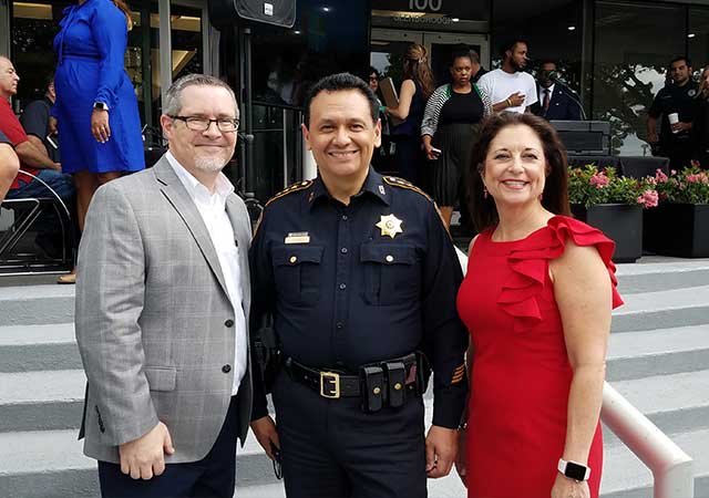 Three individuals stand on outdoor steps, smiling at the camera. A man in a suit, a uniformed officer, and a woman in a red dress are pictured with a crowd of people mingling in the background.