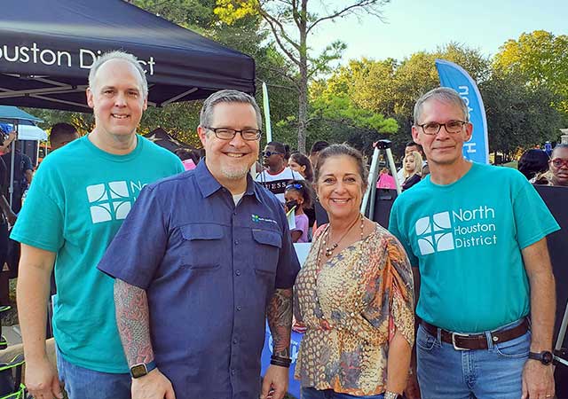 Four smiling individuals pose outdoors in front of a North Houston District tent and banner. Two men wear matching turquoise North Houston District shirts, while a woman and another man stand alongside them at a community event.