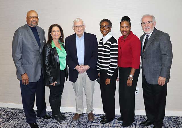 Six individuals, including three men and three women, stand together against a neutral wall background, smiling for the camera. They are dressed in business attire, with one woman wearing a green blouse and black leather blazer.