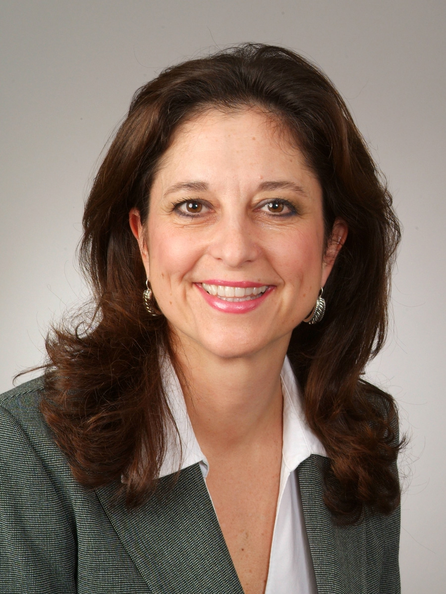 A smiling woman with shoulder-length brown hair poses in a green blouse and black blazer. She is standing against a neutral beige background.