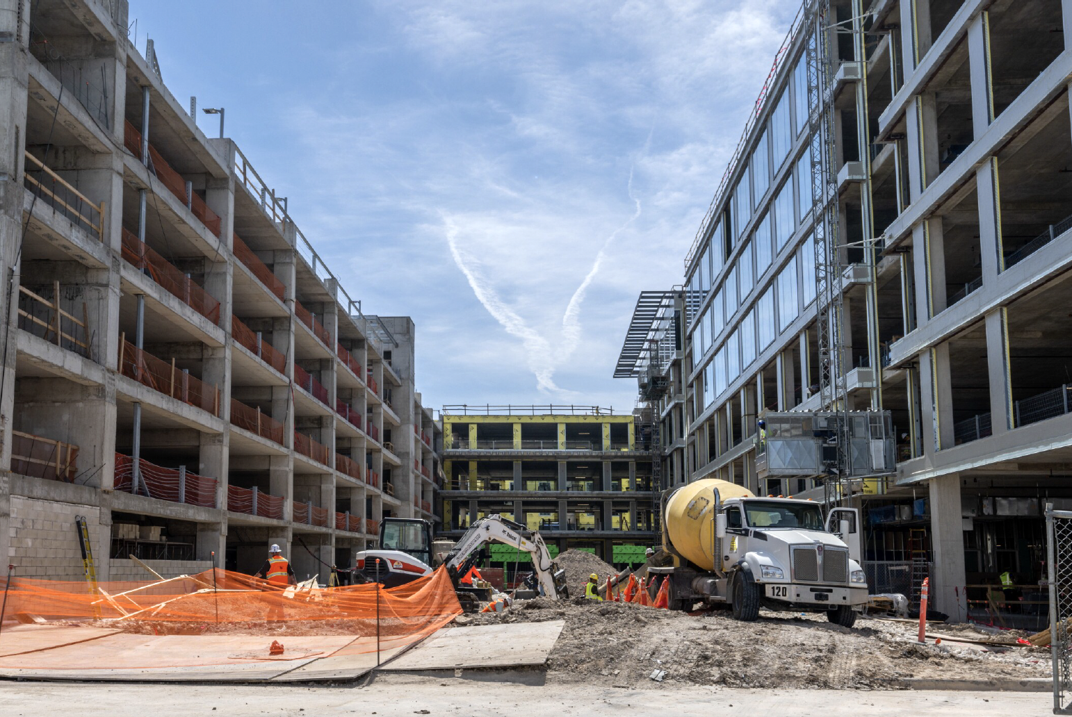 A construction site featuring two partially built multifamily buildings on either side of a central dirt pathway. Workers, equipment, and a yellow cement truck are visible, with orange safety netting and scaffolding in place. A blue sky with light clouds is above.