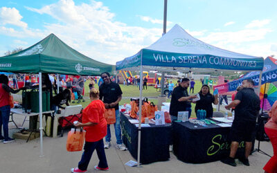 National Night Out 2025 10 Community members gather around vendor tents, including a booth labeled Villa Serena Communities, as volunteers hand out items during an outdoor neighborhood event with inflatable activities visible in the background.