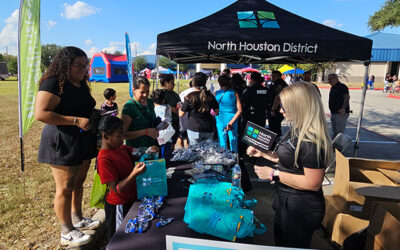 National Night Out 2025 11 Families and children line up at a North Houston District tent as staff distribute branded items across a table during a busy outdoor community event on a sunny day.