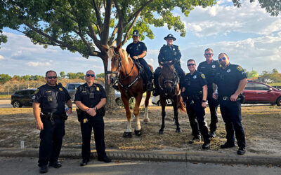 National Night Out 2025 2 Uniformed law enforcement officers stand outdoors beneath a tree, including two mounted officers on horseback, posing together at a community event on a sunny day with parked cars in the background.