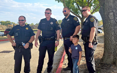 National Night Out 2025 3 Four uniformed police officers stand smiling beside a young child who poses with them on a sidewalk near a tree, creating a friendly moment during a neighborhood community event.