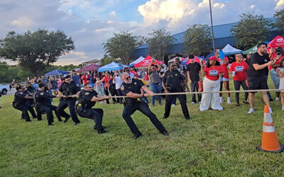 National Night Out 2025 Uniformed law enforcement officers participate in a tug-of-war game on a grassy field while a crowd of spectators watches and cheers at an outdoor community celebration.