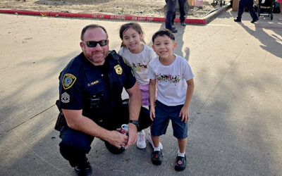 National Night Out 2025 5 A uniformed police officer kneels beside two young children on a paved area, smiling for a photo together during an outdoor community event with people and activity visible behind them.