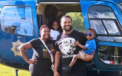 National Night Out 2025 6 A family poses smiling in front of a blue law enforcement helicopter, with two adults and two children standing near the open cabin door during an outdoor community event.