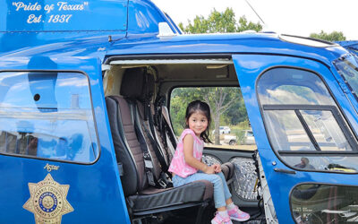 National Night Out 2025 7 A young child sits smiling inside the open cabin of a blue law enforcement helicopter, with visible seats and equipment, during a community event held outdoors.