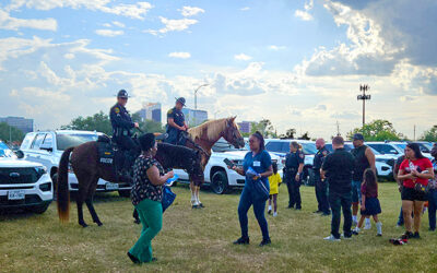 National Night Out 2025 9 Community members gather on a grassy area as mounted law enforcement officers on horseback interact with families, with patrol vehicles and city buildings visible in the background.