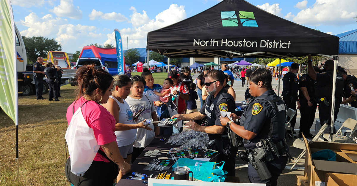 National Night Out 2025 Newsroom Graphic Uniformed law enforcement officers and North Houston District representatives engage with families at a tent, handing out safety materials and giveaways during a crowded community event.