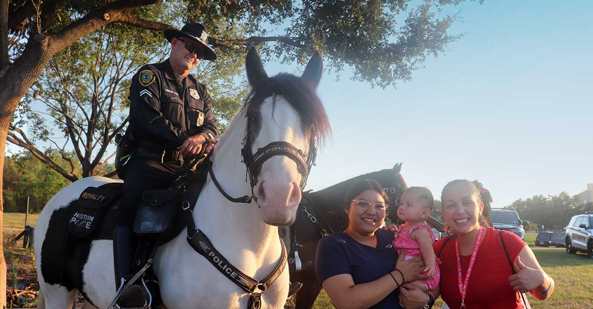 Two women are standing next to a mounted police officer. One woman in a navy outfit is holding a baby in a pink outfit. The other woman is giving a thumbs-up while smiling at the camera. The officer is seated on a large, white police horse. They are outside with trees and police cars in the background.