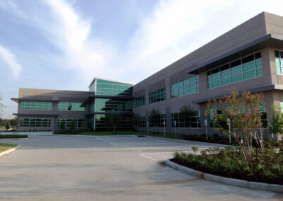 Modern mid-rise office building with glass windows, landscaped grounds, and a wide entry drive under a partly cloudy sky.