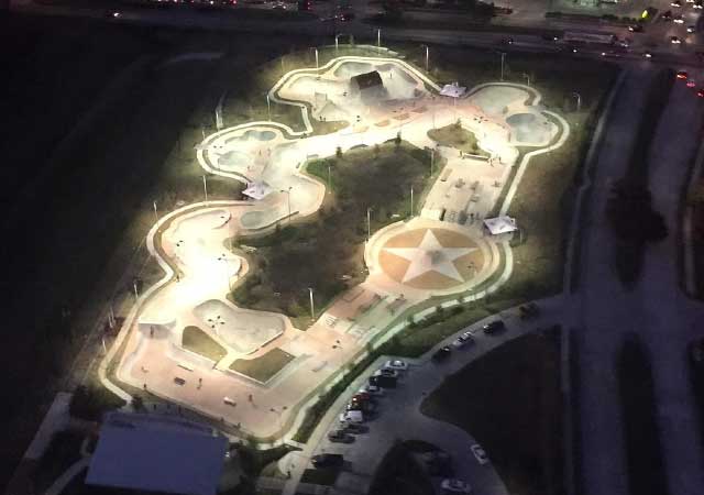 An aerial nighttime view of the North Houston Skate Park shows illuminated skate bowls, paths, and lighting arranged in an organic layout surrounded by streets and parked vehicles.