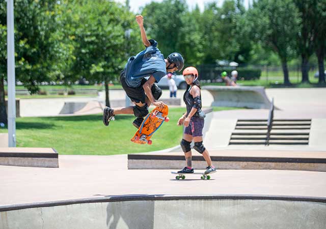 A skateboarder wearing a helmet performs a jump above a concrete bowl at the North Houston Skate Park, while another skater watches nearby on the skate plaza.