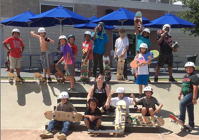 A group of children wearing helmets pose together with skateboards at the North Houston Skate Park, smiling and standing along the edge of a skate bowl during a group activity.