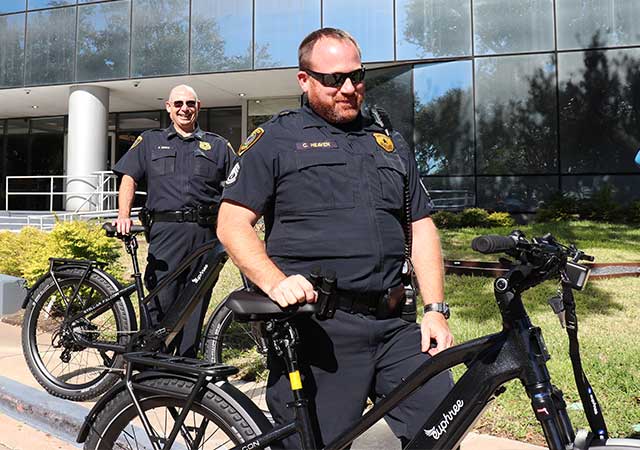 Two uniformed officers stand with black e-bikes in front of a glass building. One officer adjusts the bike seat while the other smiles. A sunny green lawn surrounds them.