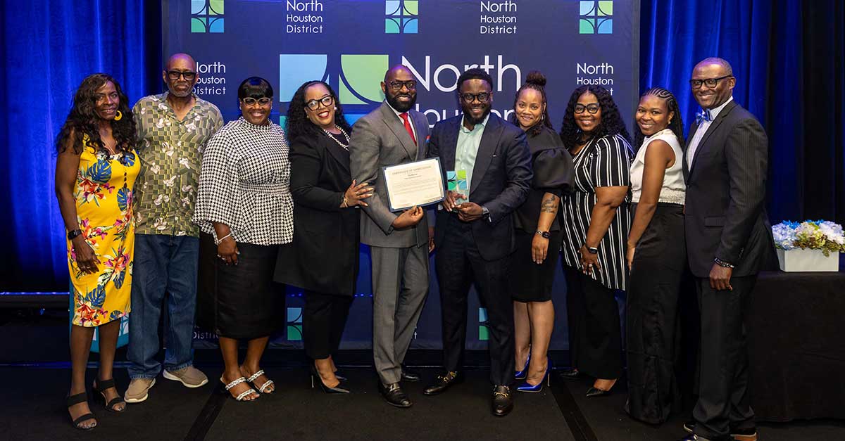 A group of 11 people stands smiling in front of a North Houston District backdrop. Two men in the center hold an award and certificate, with the group dressed in professional and semi-formal attire on a stage with blue lighting.