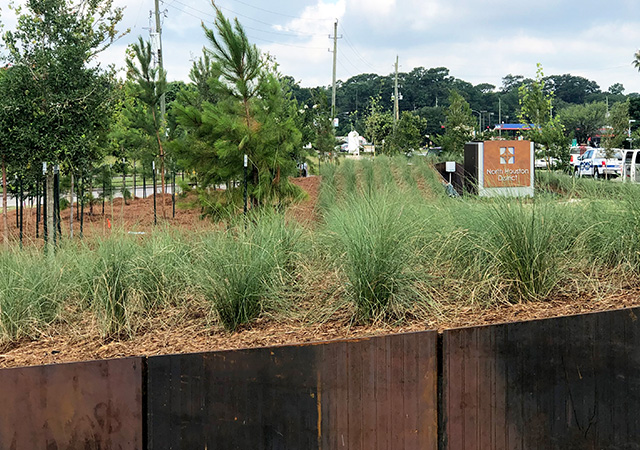 A landscaped area with tall grasses and young trees is bordered by a rust-colored steel barrier. A "North Houston District" monument sign stands in the background near a parking lot.