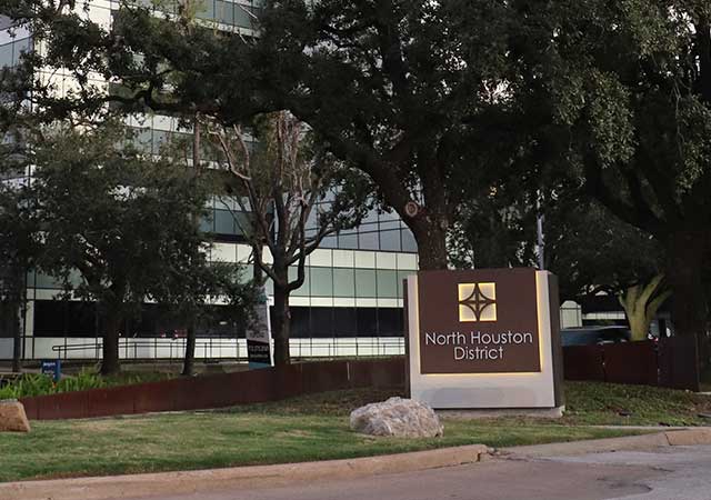 A North Houston District monument sign is framed by lush greenery and mature oak trees. A modern glass office building and a road are visible in the background.