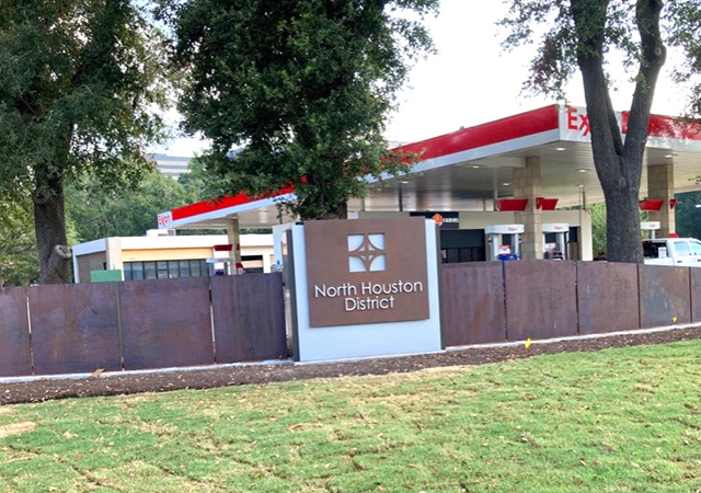 A North Houston District monument sign stands on a grassy area with a rust-colored steel wall in the background. Tall trees surround the site, and a gas station with a red canopy is visible behind the trees.