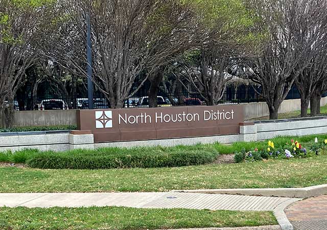 A long North Houston District monument sign rests on a landscaped area with green shrubs, colorful flowers, and a stone base. A row of trimmed trees lines the background near a fence. A paved walkway curves in the foreground.