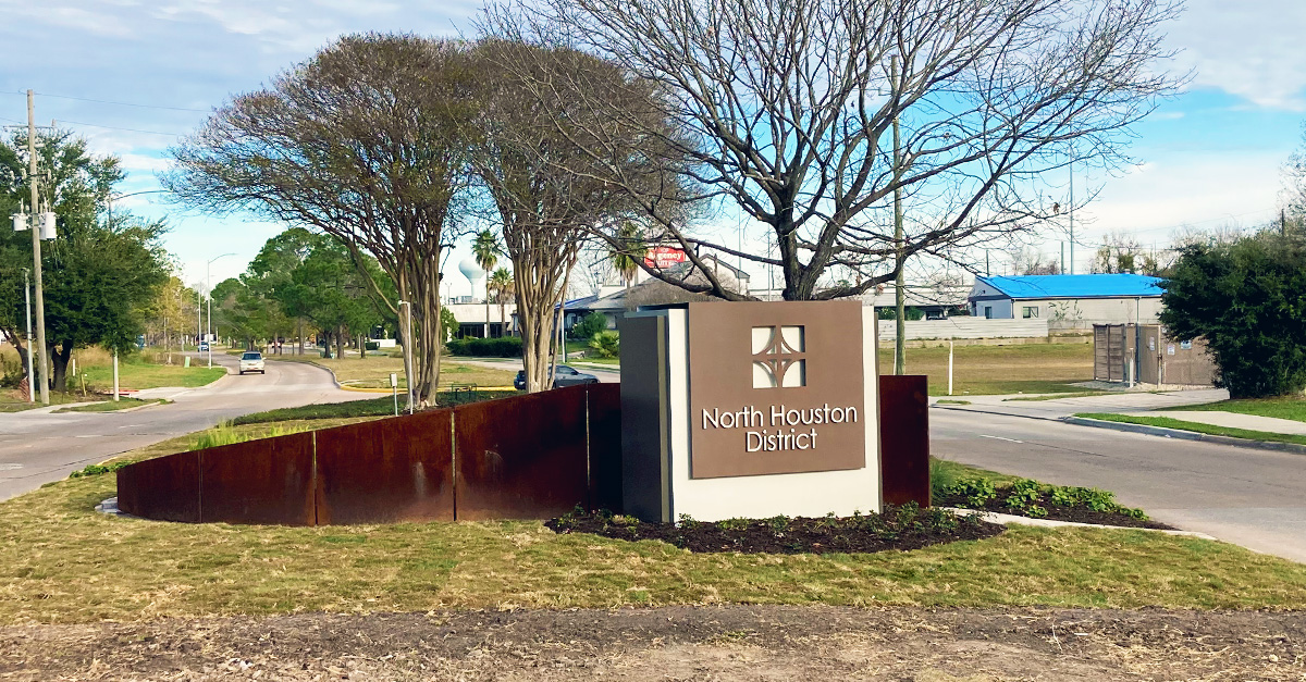North Houston District gateway monument sign installed in a landscaped median near a roadway intersection. The sign features the District logo, decorative steel accents, and surrounding trees and greenery.