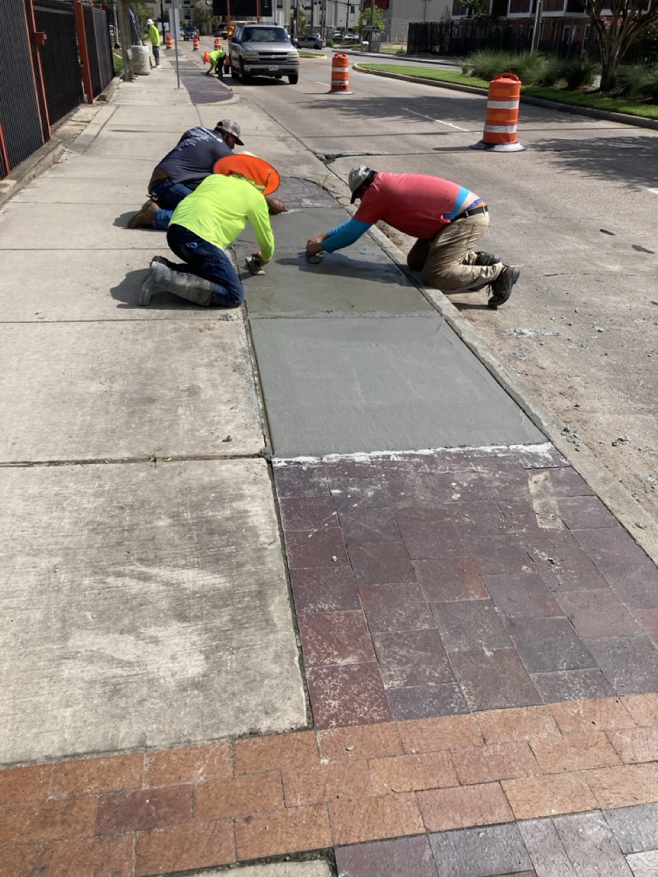 A group of construction workers kneel and smooth out wet concrete on a sidewalk segment, with orange cones and a vehicle in the background.