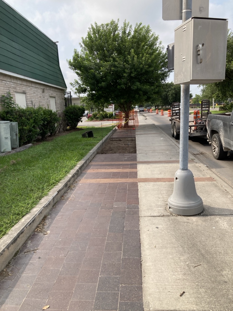 A partially excavated sidewalk lined with brick pavers and surrounded by greenery. Construction barriers and cones are visible ahead, marking the work zone.