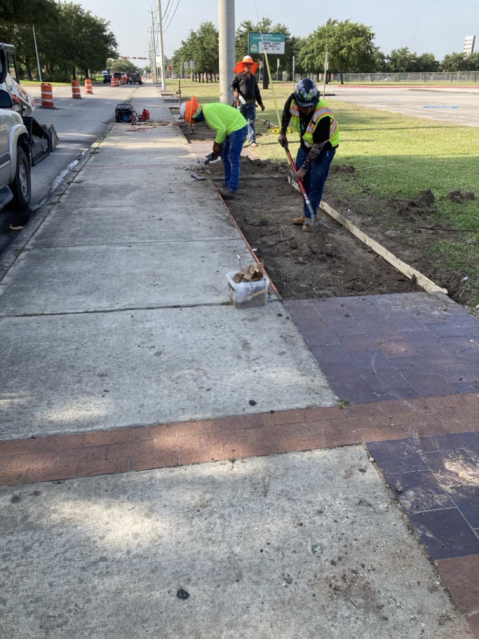 Workers use tools to dig and prepare the ground for new sidewalk sections, with orange cones marking the construction area along the street.