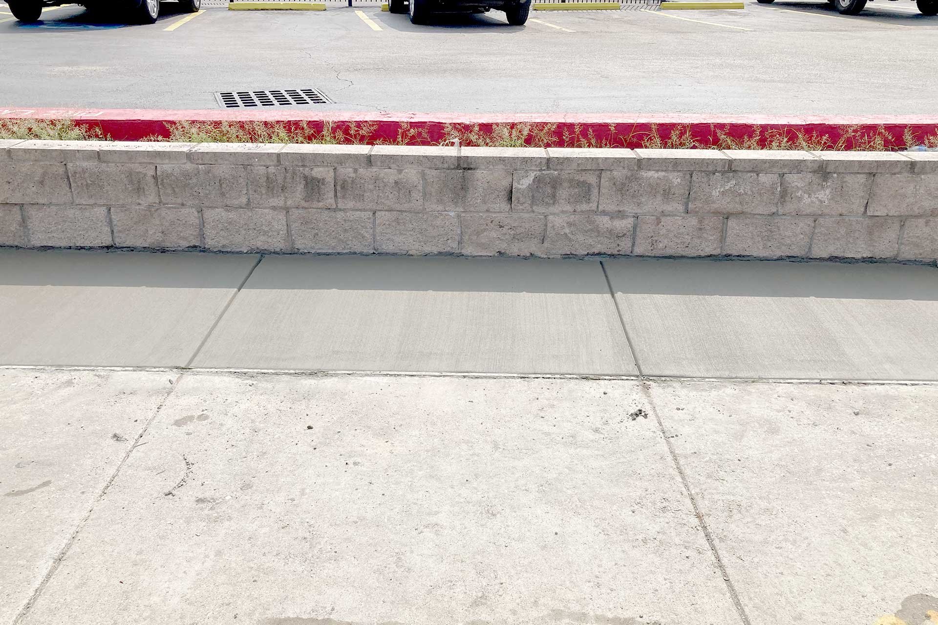 Newly poured concrete sidewalk stretches along a parking lot with a retaining wall and a red-painted curb.