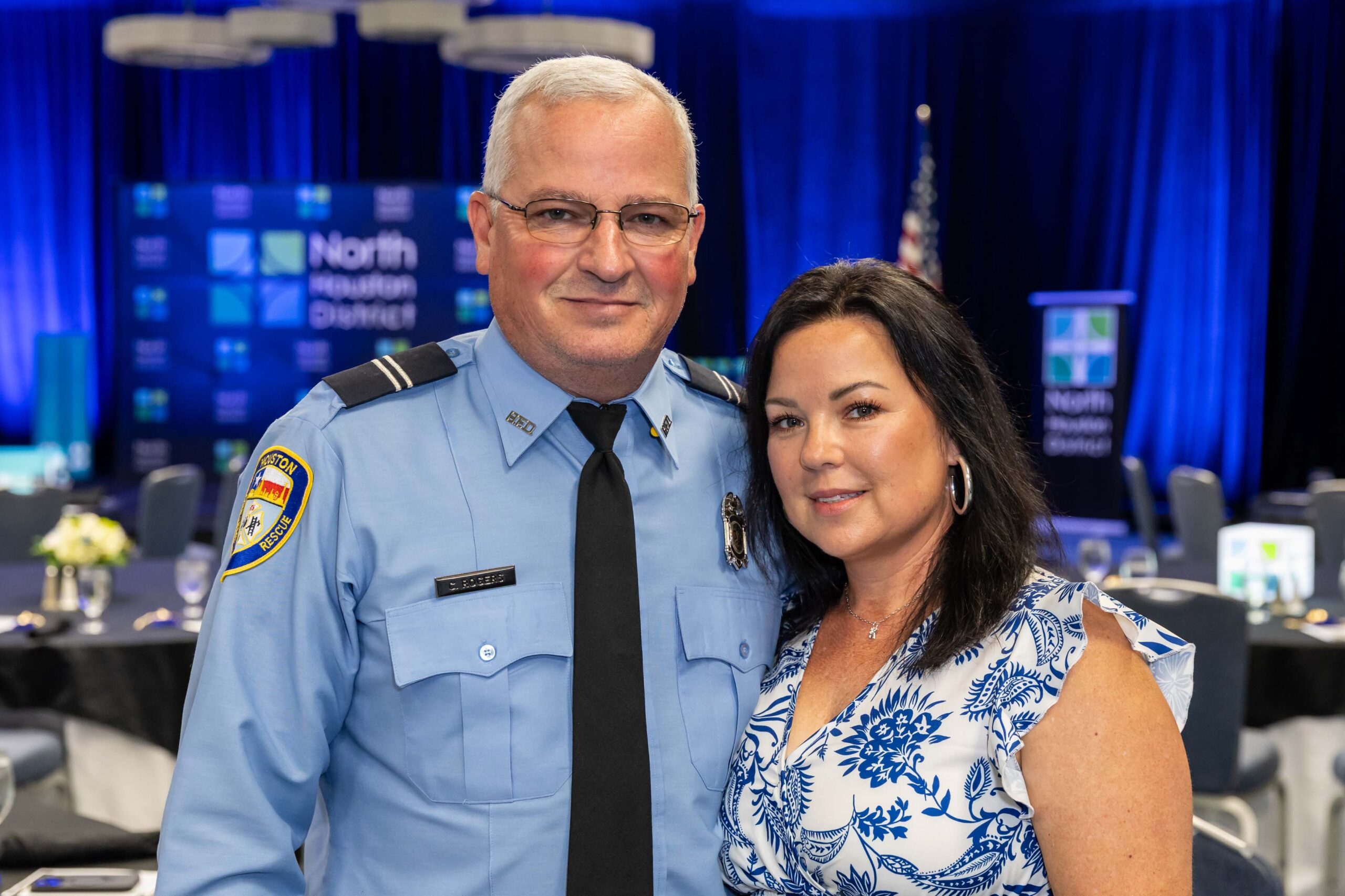 A uniformed HFD officer and a woman pose in front of a blue-lit stage and branded backdrop.